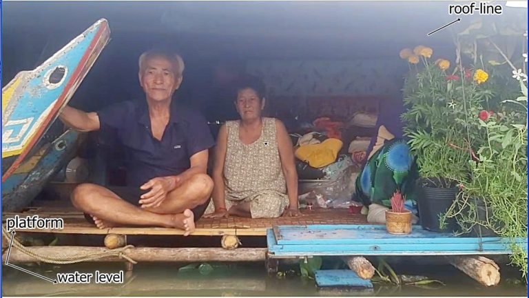 An annotated photo of an elderly couple sitting on a raised platform inside their flooded home, with labels showing the roof-line, platform, and water level