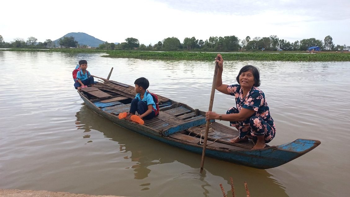 A grandmother smiling as she steers a wooden boat carrying her two grandchildren in school uniforms across the Tonle Sap