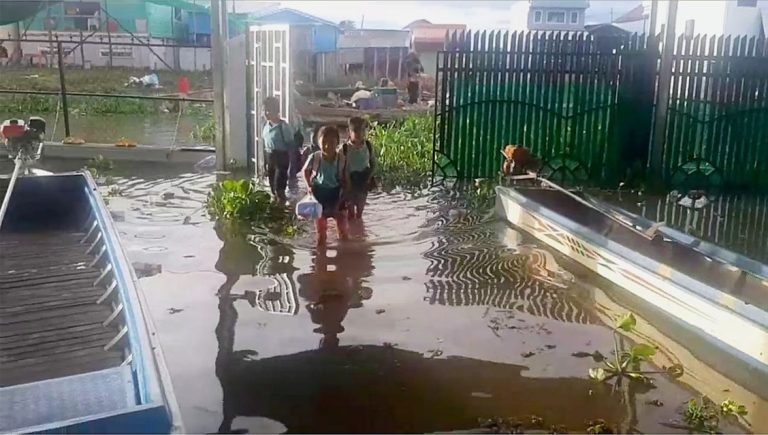 Three young students with backpacks wading through floodwater between fences in Tri An