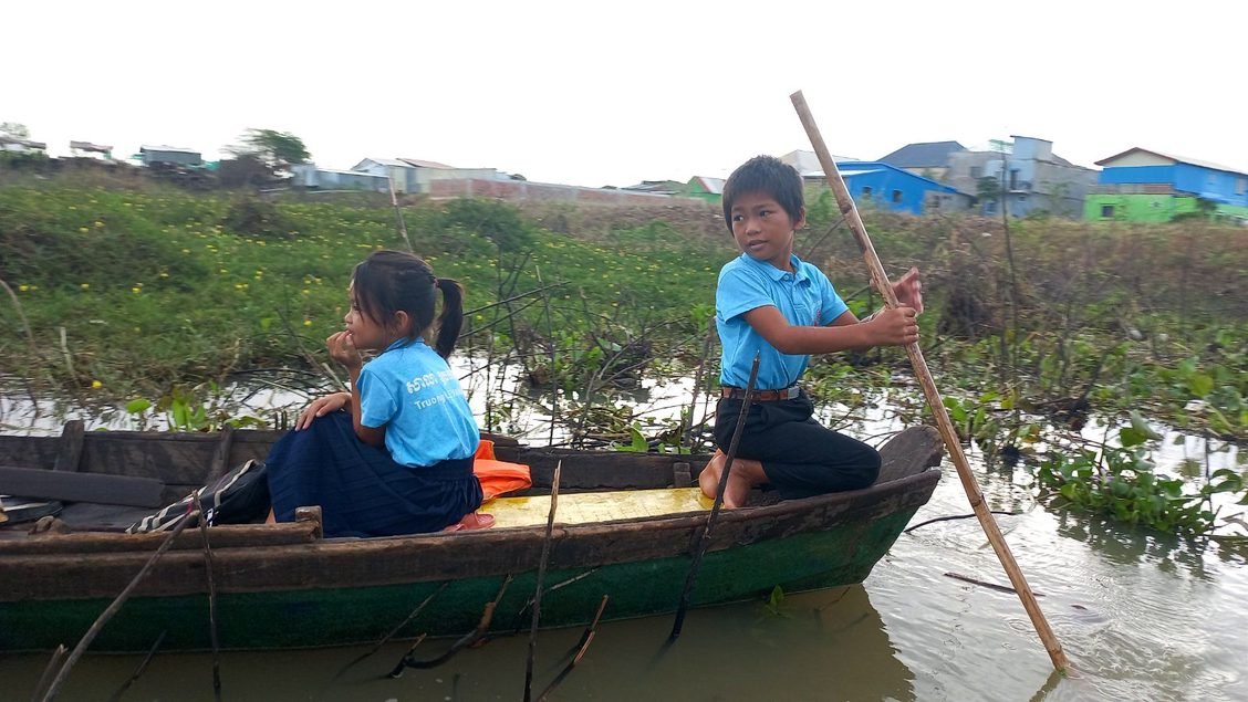 An older brother standing with a long pole in a small boat with his younger sister, both in school uniforms