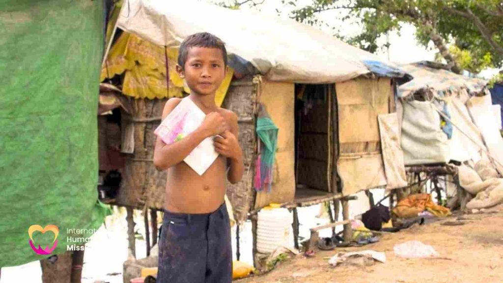 A young shirtless boy clutching a notebook to his chest, standing barefoot in front of a makeshift tarp-and-bamboo shelter where his family lives