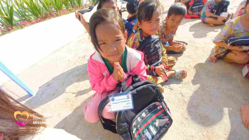 Khanh, an 8-year-old girl in a pink jacket, kneeling on the ground with her hands clasped in greeting and her new backpack on her shoulders