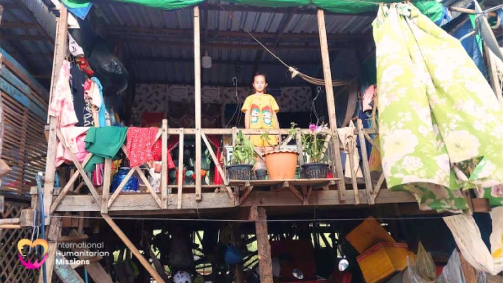 A child standing on the wooden balcony of a stilt house in Kampong Chhnang, with laundry and household items hanging around