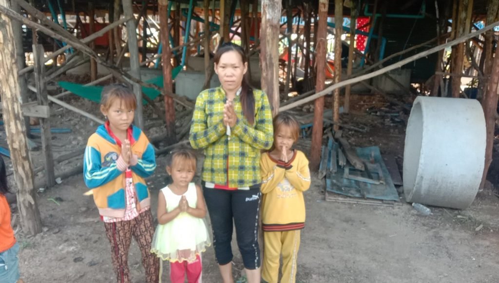 Mai standing with her three children, all with hands clasped in greeting, in front of the new toilet inside their home